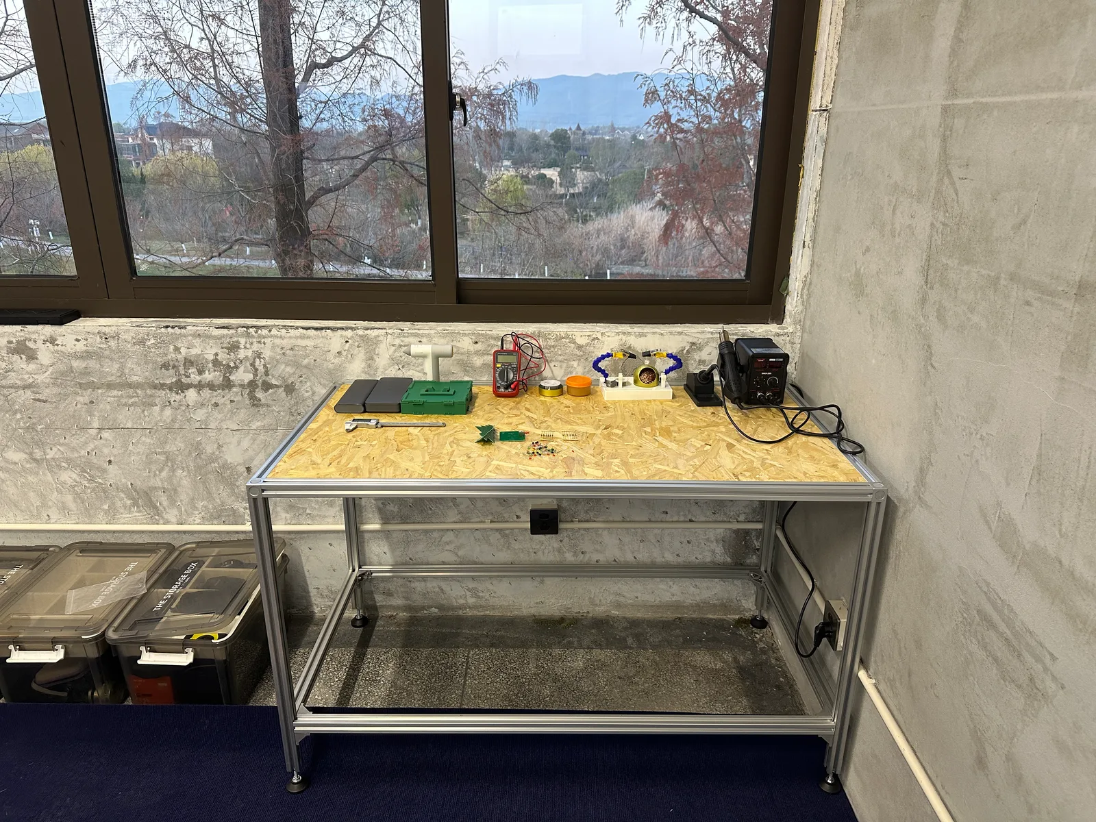A workbench with an aluminium frame and a board top under a window, with tools and a soldering station on it