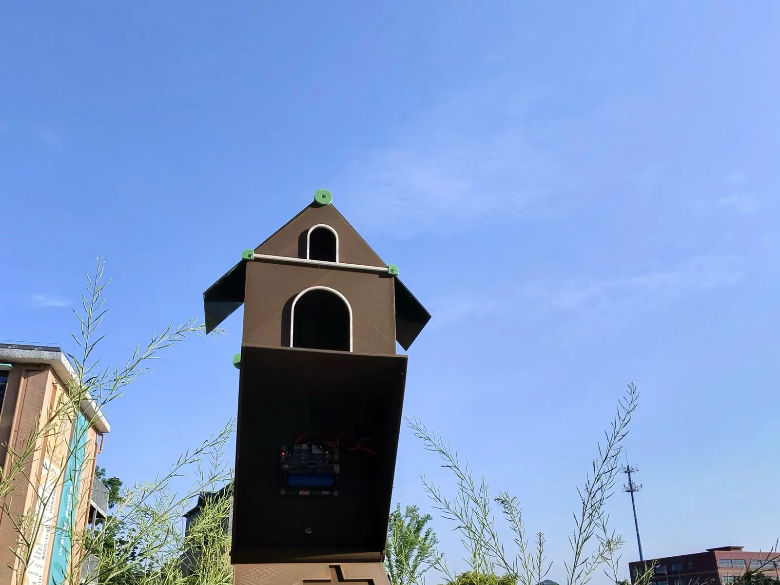 A dark printed birdhouse with a solar panel on a post above wooden vegetable beds, against a blue sky