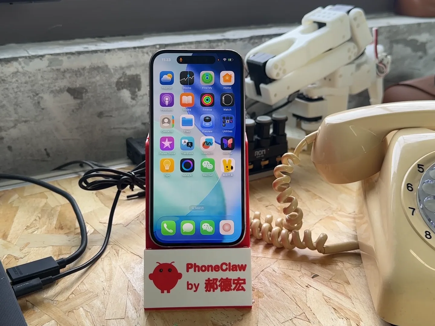 A phone standing in a red 3D-printed dock labelled PhoneClaw on a workbench, beside a cream rotary telephone and a white printed robot arm