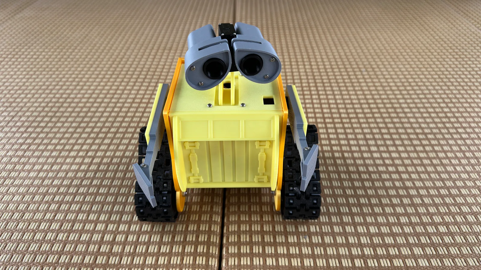A small yellow 3D-printed robot with tracks and two round camera eyes on a woven mat