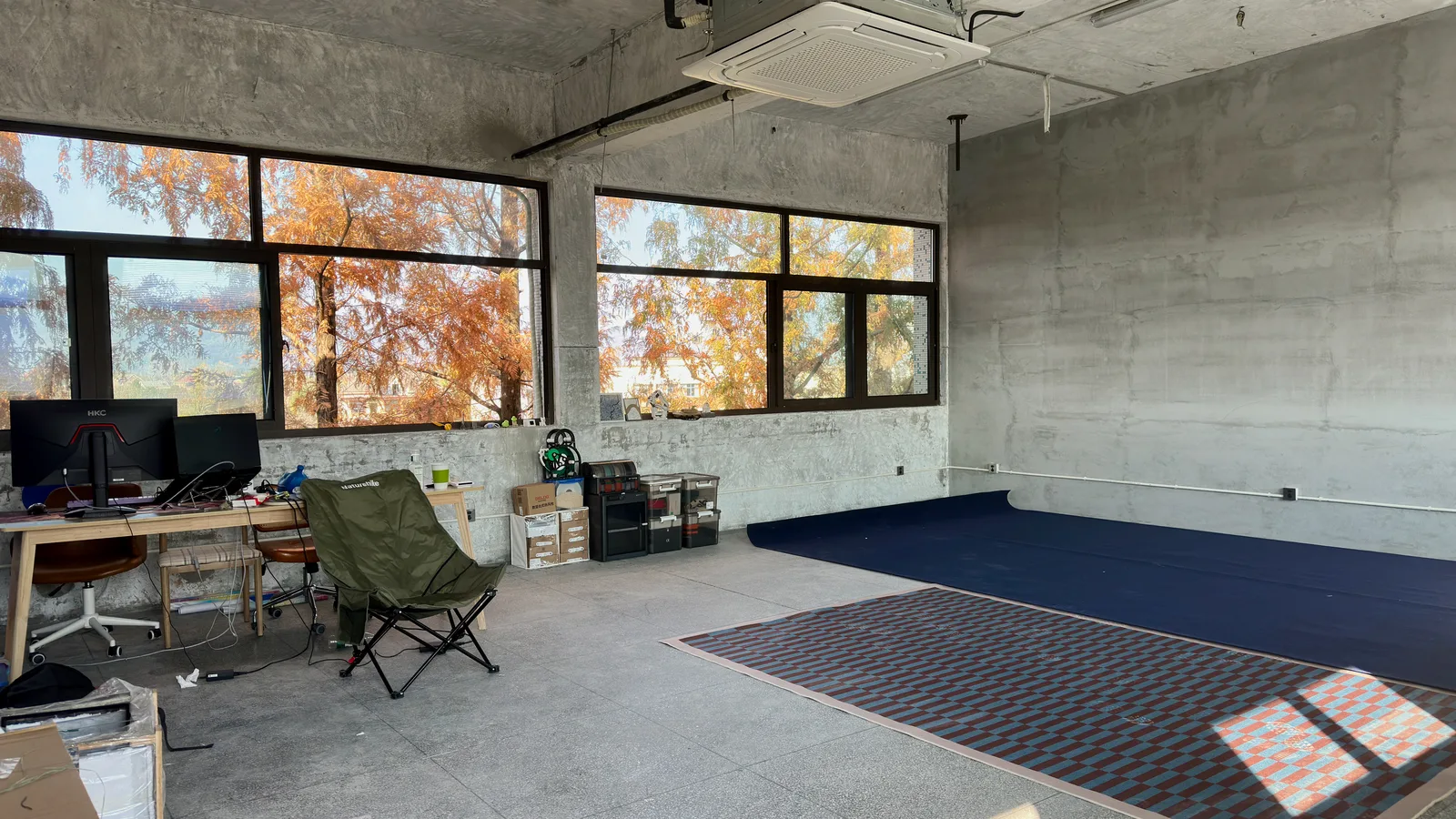 A nearly empty concrete classroom with a camping chair, a desk in one corner and autumn trees in the windows