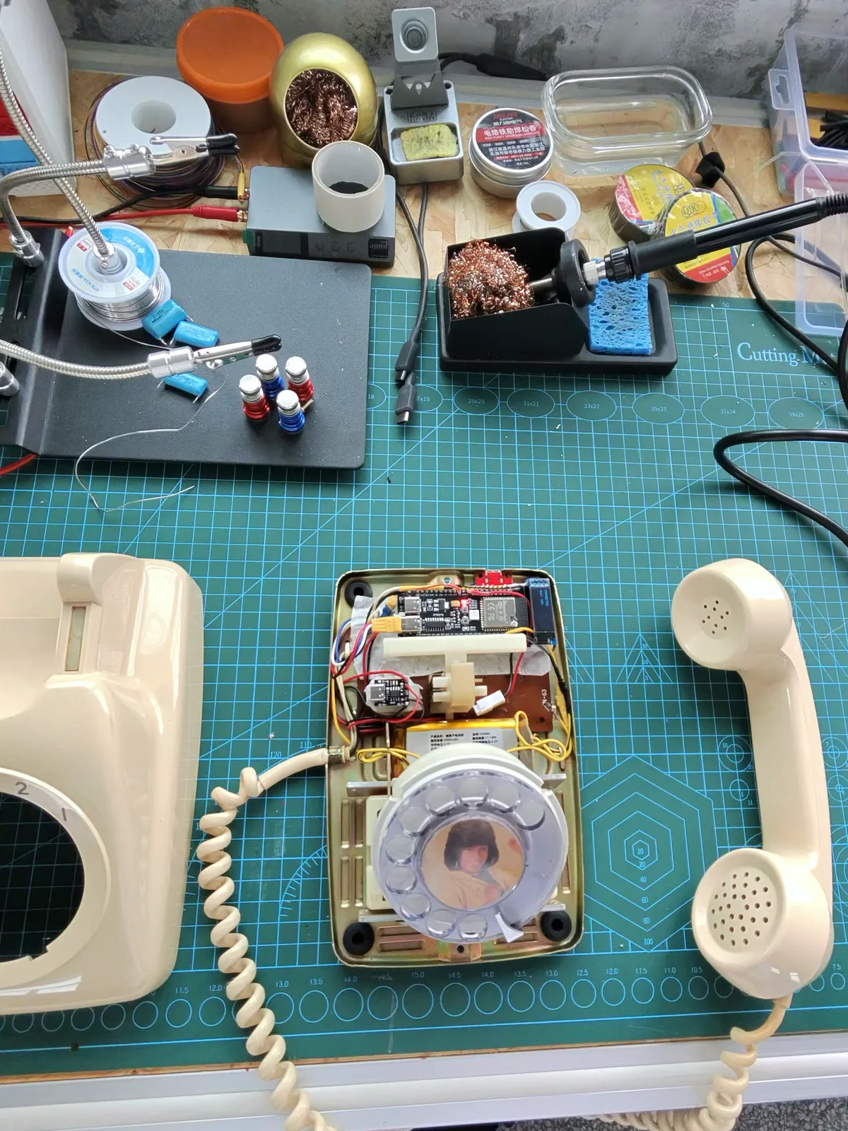 A cream rotary telephone taken apart on a cutting mat, its dial and handset laid out beside a soldering iron
