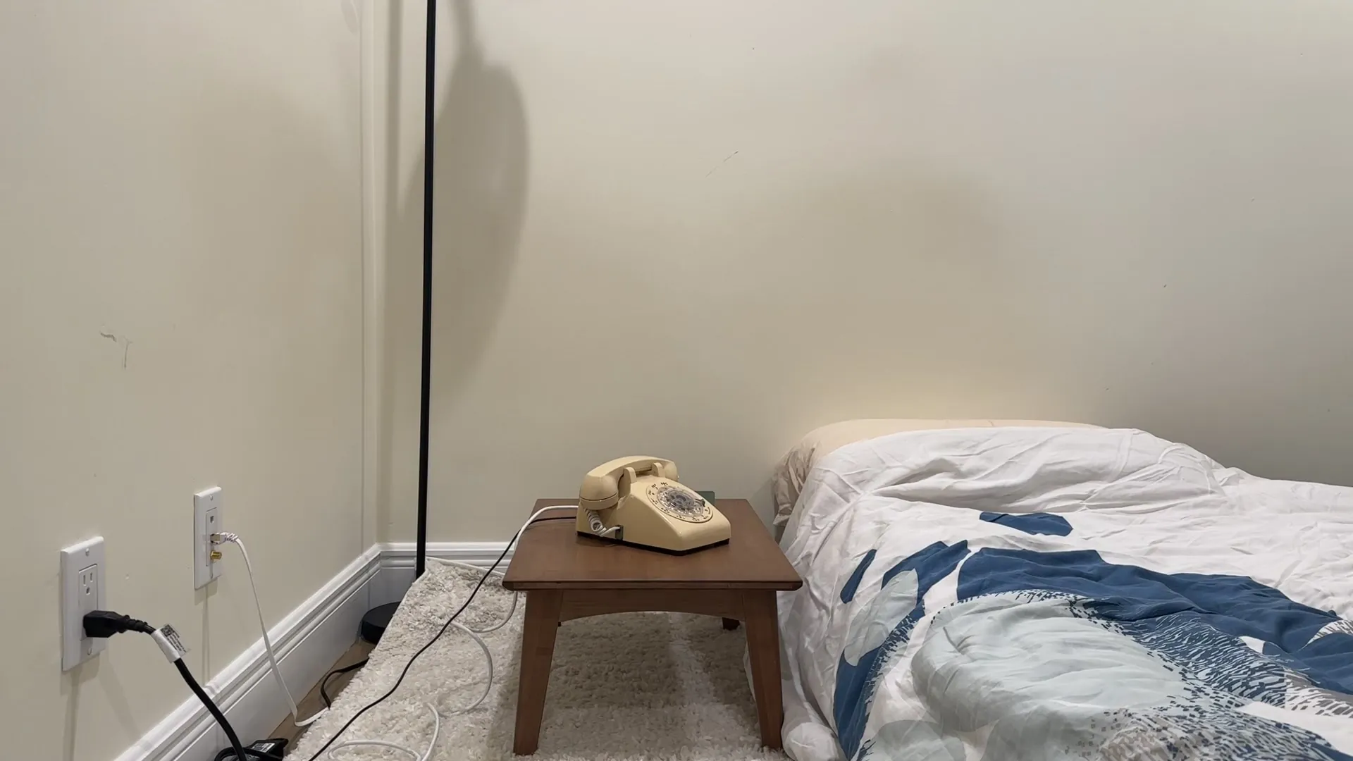 The cream rotary telephone on a low wooden table beside a bed, against a plain wall
