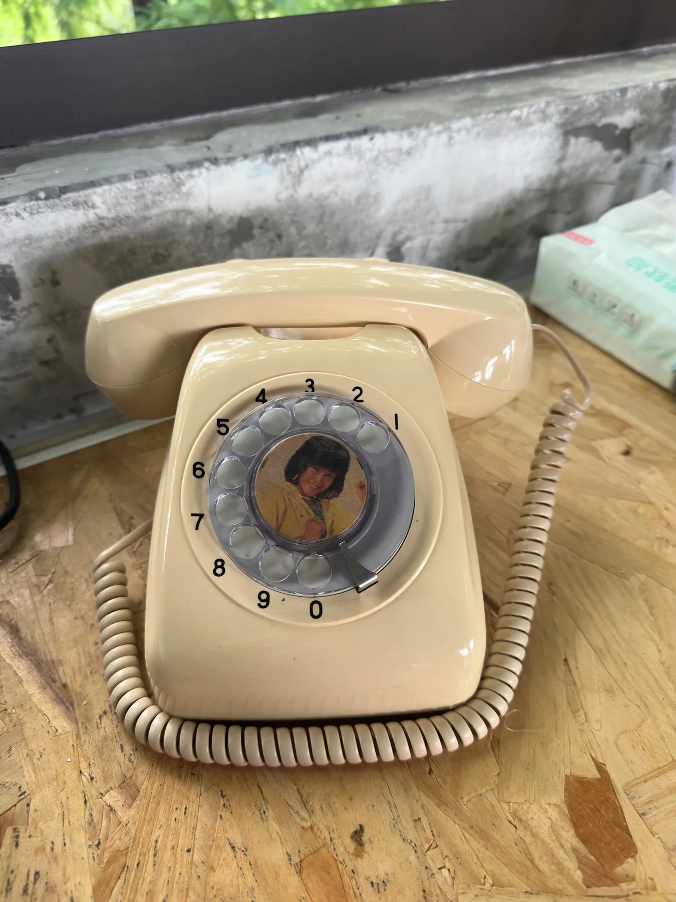 A cream rotary telephone with a coiled cord on a plywood desk; a small portrait of a woman sits in the center of the dial