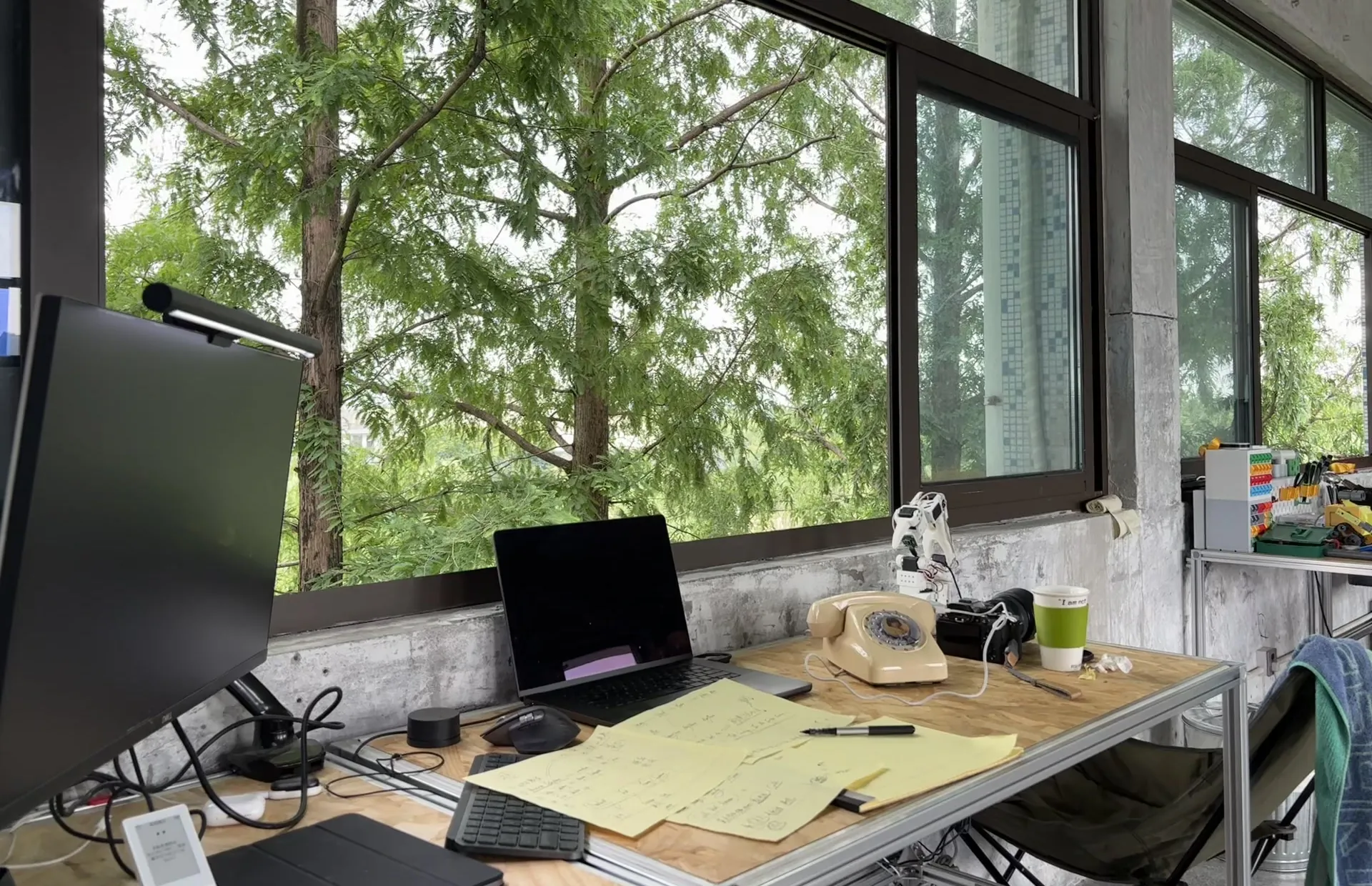 A cream rotary telephone on a plywood desk beside a laptop and yellow notes, under tall windows full of trees